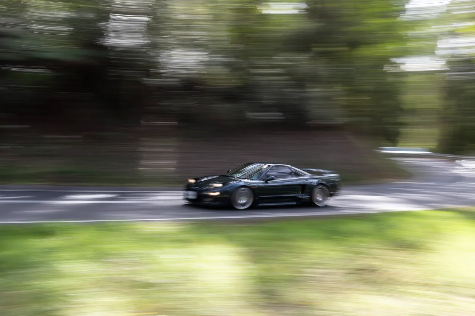 Black sports car with headlights on driving through a tree-lined road with motion blur surrounding the vehicle.