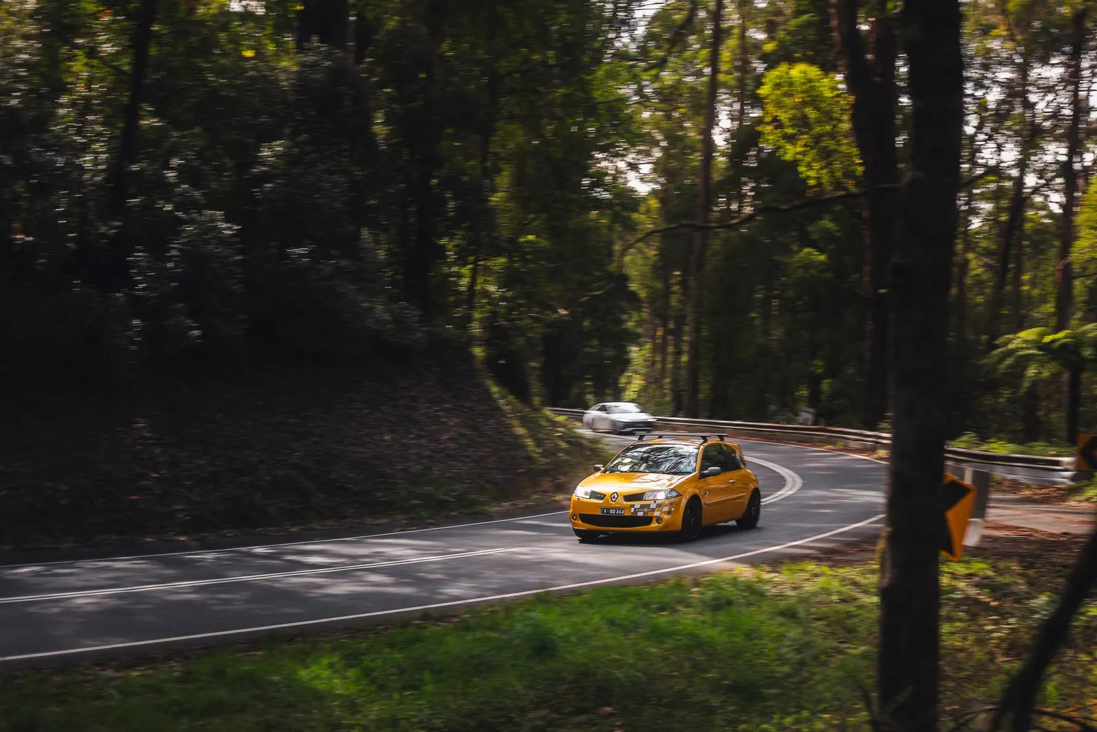 A yellow compact car cornering through a wooded road with a white vehicle visible behind it.