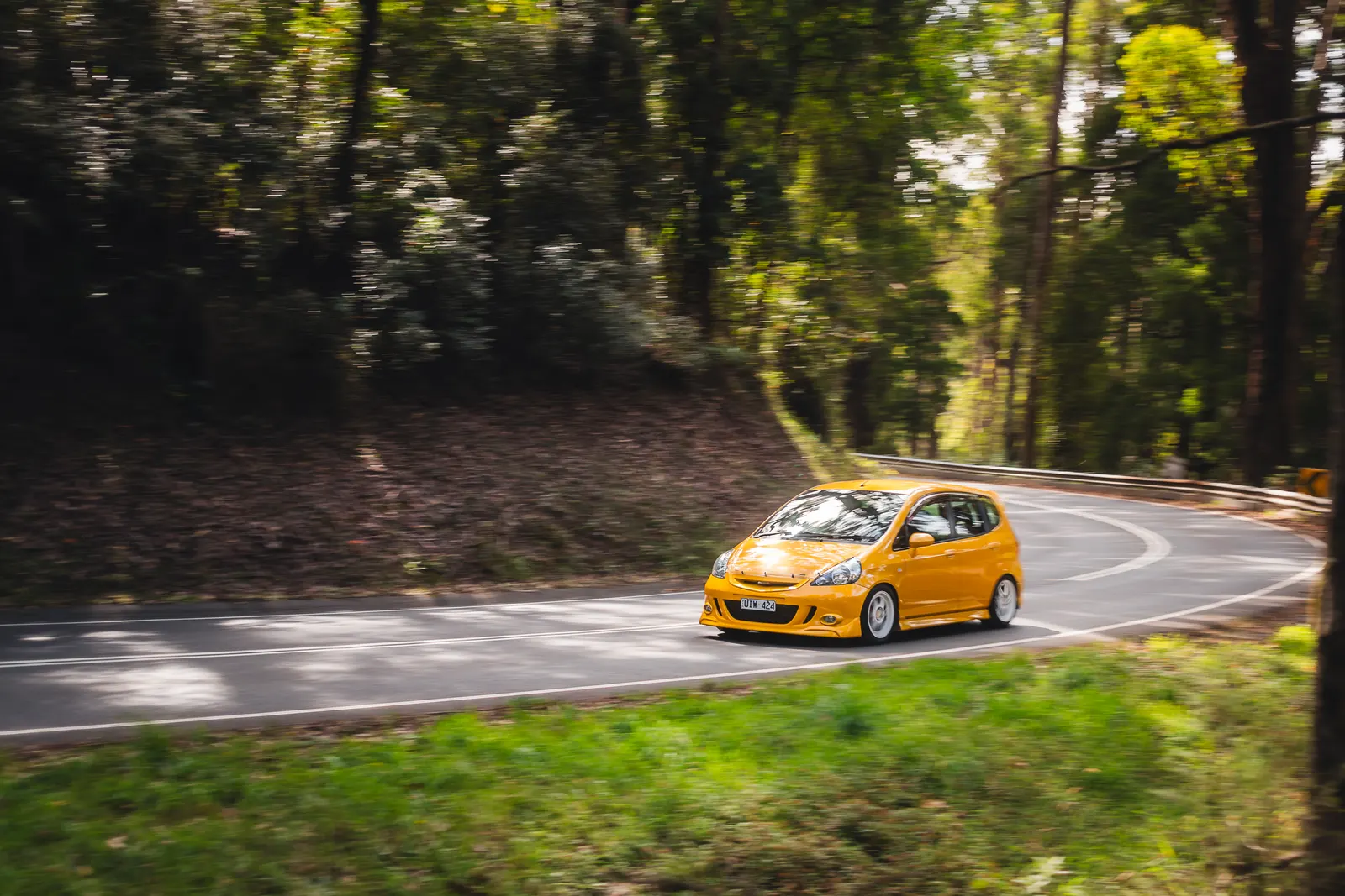 Yellow hatchback driving through a tree-lined road with dappled sunlight and motion blur.